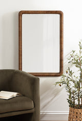 Rectangular wall mirror with a dark brown beaded wood frame, reflecting light from a window, positioned above olive-green upholstered sofa and woven basket with greenery.