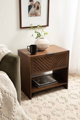 Dark wood side table with diagonal slat front panel, open lower shelf holding books, topped with a white ribbed vase and black mug, beside green sofa and cream textured rug.