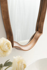 Curved wooden-framed wall mirror with flowing scalloped edges, shown close-up above white flowers and a smooth white surface.