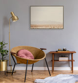 Modern round tan leather chair with black metal legs and a pink round cushion, next to a wooden side table and brass floor lamp against a gray wall with a desert landscape painting.