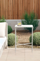 Slim white metal side table with rectangular top and crossed legs holding a glass of water with lemon, positioned outdoors on tiled patio near wicker sofa and plants.