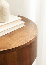 Close-up of a round wooden side table with smooth curved edges and rich brown finish, holding white hardcover books and a textured beige vase against a light curtain background.