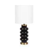 Table lamp with stacked black rounded resin rings as base and a white cylindrical fabric lampshade, accented with brass metal elements at top and bottom.