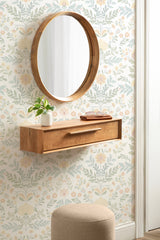 Rectangular floating wooden console table with a single drawer beneath a round wooden-framed mirror on floral wallpaper, with a beige upholstered stool below.