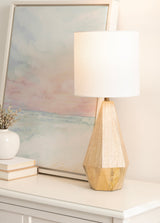 Table lamp with a faceted natural wood base and a white cylindrical shade, positioned on a white surface beside an abstract pastel seascape painting.