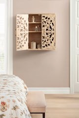 Wall-mounted wooden cabinet with intricately carved floral designs on double doors, partially open to reveal two shelves holding small decorative items, against a beige wall.