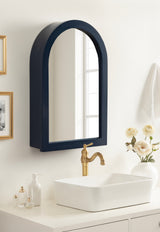 Arched wall mirror with a deep navy blue frame above a white rectangular vessel sink and brass faucet on a white countertop with cream-colored flowers and glass bottles.