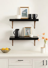 Two black floating shelves with wooden brackets mounted on a white wall, decorated with framed photos, black mugs, a black pitcher, and white ceramic containers, above white cabinetry with black handles.