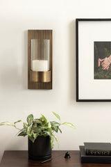 Vertical metal wall sconce with ribbed rectangular backplate, cylindrical glass candle holder, and a white candle, mounted above a plant and books on a dark surface.