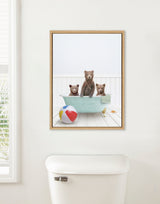 Framed canvas showing three brown bear cubs sitting and standing in a light blue clawfoot bathtub on whitewashed wooden floor beside a colorful beach ball and starfish.