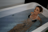 Woman with eyes closed relaxing in a white rectangular acrylic tub filled with clear water, wearing a black bikini with white straps.