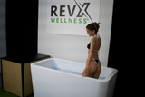 Woman in black and white bikini standing in a rectangular white acrylic cold plunge tub partially filled with water inside a minimalist indoor space.