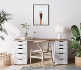 Minimalist home office with wooden desk featuring white drawer units, a light wood chair, potted plants, a table lamp, and framed flamingo portrait on white wall.