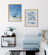 Two framed artworks on a white wall above a light blue sectional sofa; left is an abstract seaside scene with three boats in blue and beige tones, right features six stylized blue fish on beige background.
