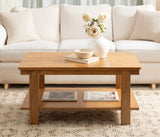 Rectangular wooden coffee table with a lower shelf holding magazines, topped with a white vase of cream flowers and a matching bowl, set on a patterned rug in front of a cream sofa.