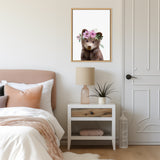 Framed canvas of a brown bear cub wearing a purple flower crown, hanging above a nightstand with a beige lamp and a small potted plant in a minimalist bedroom.