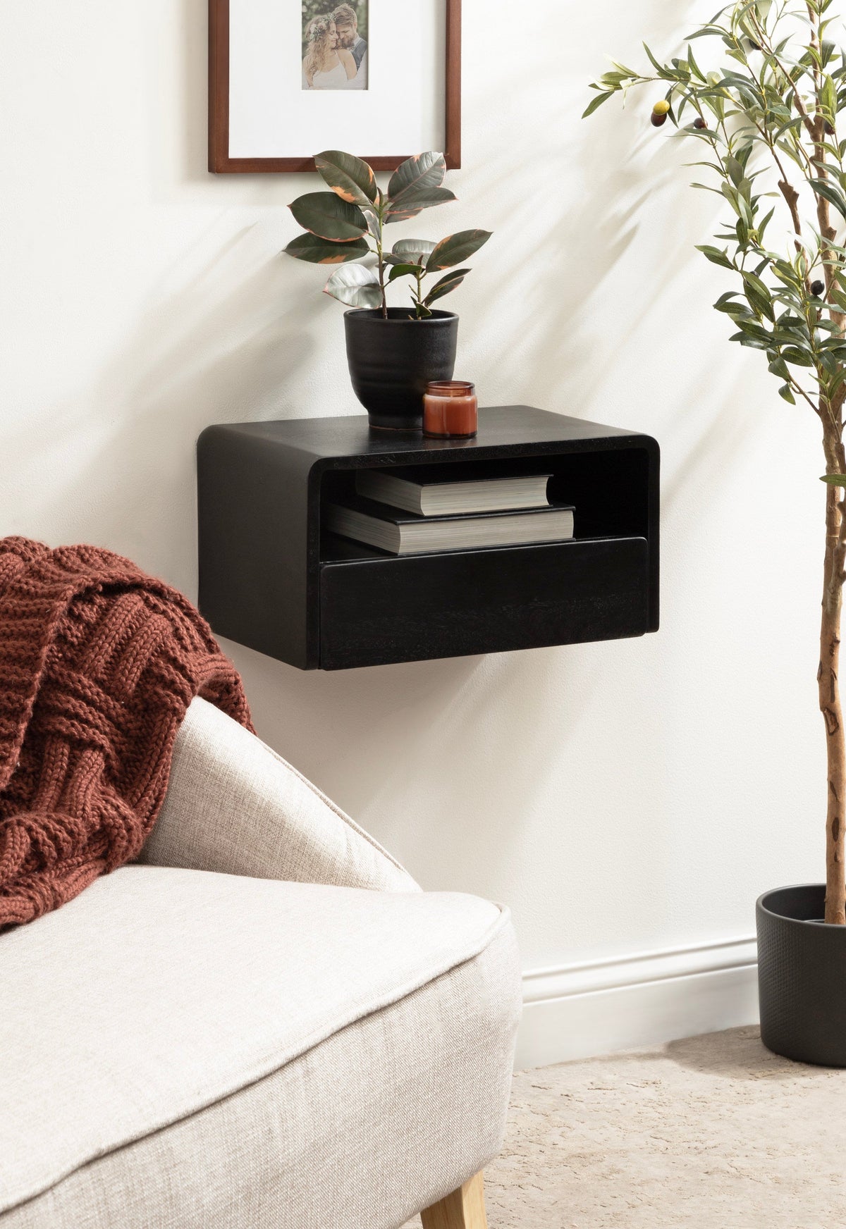 Black floating side table with rounded edges mounted on a white wall, holding a small potted plant and a candle, above a beige sofa with a rust-colored knit blanket.
