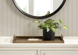 Rectangular brass-colored metal tray with raised edges and handles, holding a black pot with green variegated leaves and a small envelope on a white surface beneath striped wallpaper and a round mirror.