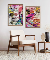 Two abstract floral canvas art pieces with bold brushstrokes of pink, purple, yellow, and black framed in gold, displayed above a mid-century style wooden armchair.