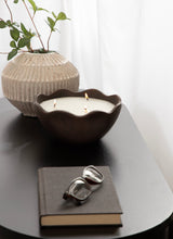 Dark brown metal bowl candle with three lit wicks and scalloped edges on a black table beside a ribbed beige ceramic vase with green leafy stems and a closed gray book with eyeglasses on top.