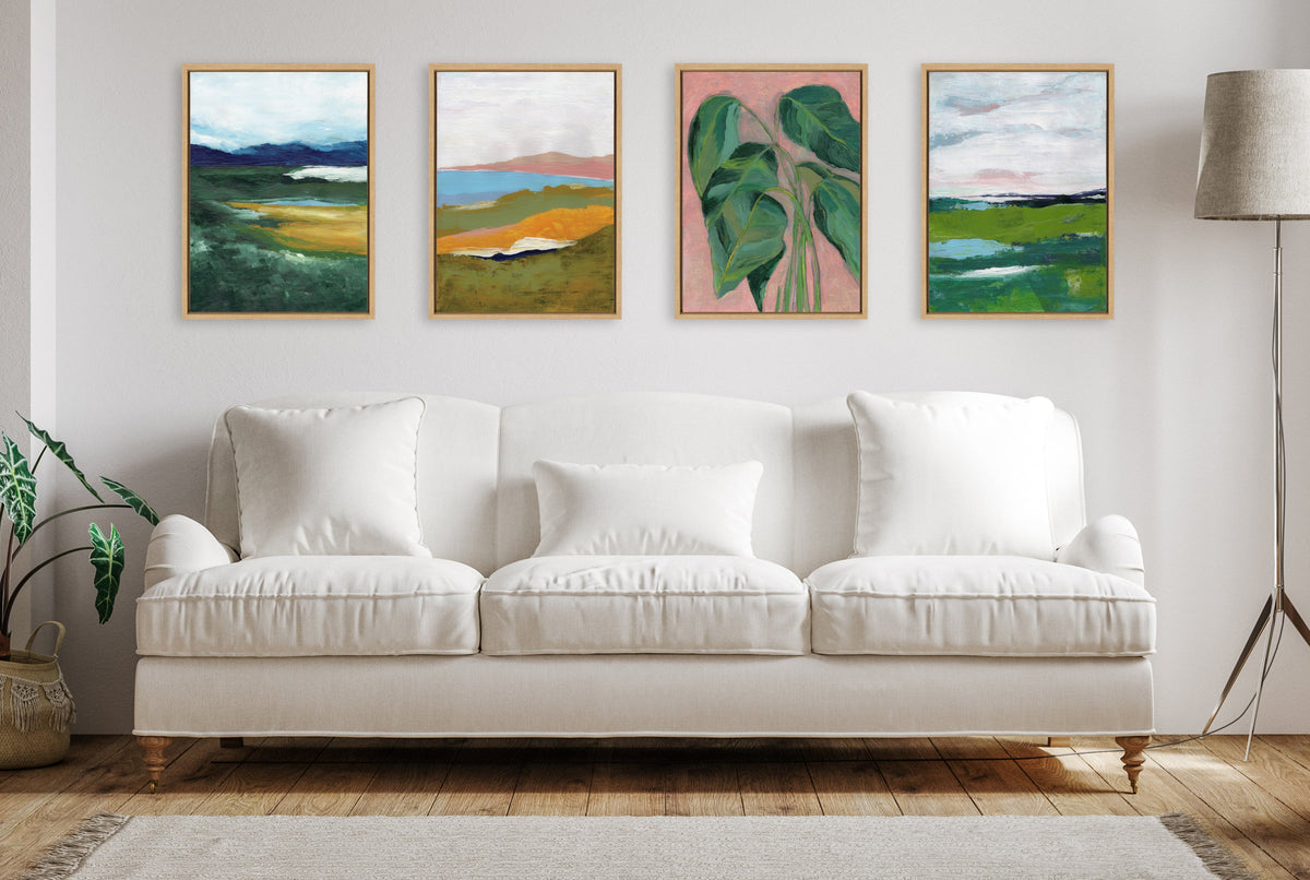 Four framed canvases with botanical and landscape paintings in green, blue, pink, and brown tones arranged above a white sofa in a minimalist living room with wooden floors and a floor lamp.