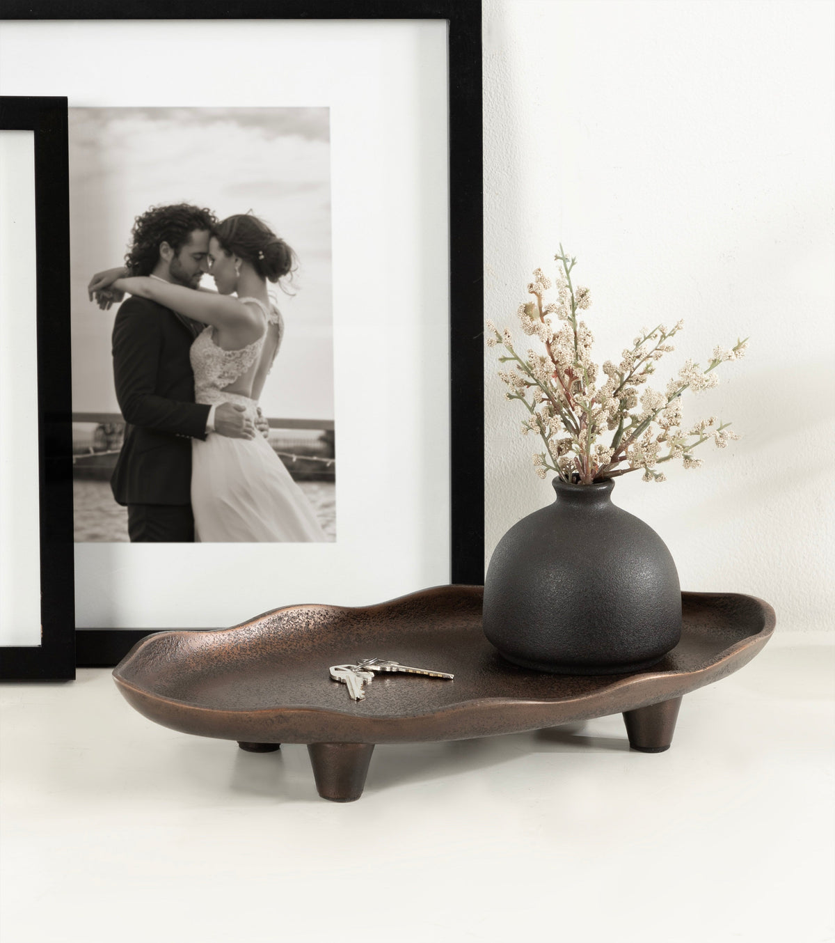 Dark brown oval tray with textured edges and four short legs, holding a matte black round vase with delicate white blossoms, set on a white surface next to framed black-and-white photo.