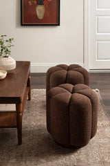 Two round, tufted ottomans upholstered in brown textured fabric, placed on a patterned rug near a wooden coffee table with a small plant and a framed floral painting on the wall.