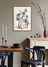 Minimalist black line drawing of three vases with flowers in a beaded wooden frame on a beige wall, above a mantel with decorative pottery and a dining table with candles.