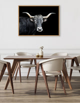 Framed canvas print of a black longhorn bull with white horns on a solid black background, hung above a modern wood dining table with cream chairs.