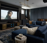 Dark blue sectional sofa with patterned pillows in a home theater room featuring a wall-mounted TV, textured area rug, and dark curtains covering a window.