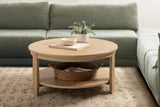 Round light wood coffee table with a lower shelf holding a woven basket, placed on a textured beige rug in front of a muted green upholstered sofa.