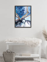Framed canvas painting of a reindeer with large antlers against a snowy blue sky, hanging above a white bench with a faux fur throw and gray handbag.