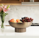 Bronze ribbed metal bowl pedestal holding peaches and red and black grapes, placed on a white kitchen counter with pink lilies in a glass vase nearby.
