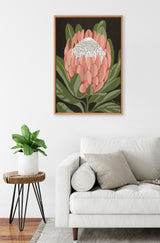 Framed canvas of a stylized Protea flower with pink petals, white center, and green leaves against a black background, hanging above a white sofa and wooden side table.