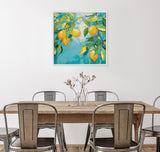 Framed canvas painting of a lemon tree branch with bright yellow lemons and green leaves against a turquoise background, hanging above a wooden dining table with metal chairs and a glass vase with twigs.