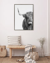 Black-and-white framed canvas of a Highland cow with long hair partially covering its face, hanging above a wooden bench with striped cushions and decor in a minimal room.