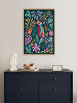 Colorful stylized parrot among various tropical leaves and flowers in bold pink, blue, green, and yellow hues, framed and hung above a dark navy dresser.