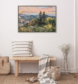 Framed landscape painting of sunlit pine trees on a rocky hill with distant blue mountains under a pastel pink and orange sky, hanging above a wooden bench with striped pillow and throw.