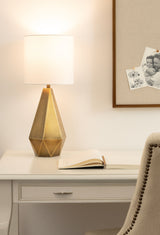 Table lamp with a faceted brass base and a white cylindrical shade on a white desk, next to an open book and a beige chair with dark nailhead trim.
