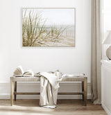 Framed canvas showing beach grass on sandy dunes with soft beige and green tones, hanging above a wooden bench with a draped white cloth in a neutral room.