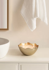 Curved matte gold metal candle bowl with three lit wicks, positioned on a white bathroom counter beneath a hanging white towel and near a round sink and mirror.