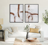 Living room with two framed abstract canvas art pieces featuring brown curved lines on a white background above a white sofa with neutral and mustard pillows.