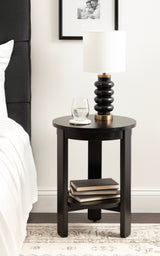 Black round wood side table with a lower shelf holding three books, topped with a black and brass lamp and a glass of water on a coaster, beside a white bed.
