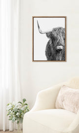Framed black-and-white portrait of a Highland cow with long shaggy hair, featuring one large horn, hanging on a white wall above a cream armchair and plant.