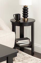 Black round wood side table with a lower shelf holding three stacked books, topped with a black ribbed lamp and a black ceramic mug beside a beige sofa on a patterned rug.