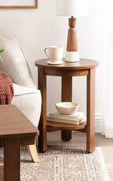 Round wooden side table with a lower shelf holding books and a bowl, topped with a wooden and white lamp and a ceramic mug, beside a beige upholstered chair.