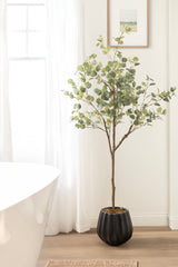 Tall artificial eucalyptus tree with green round leaves in a black ribbed planter, positioned in a bright room with light wood flooring and sheer white curtains.