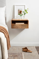 Floating side table with rounded edges and an open shelf in natural wood, mounted on a white wall next to a bed with beige and white bedding.