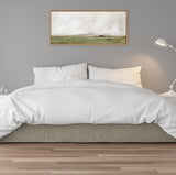 Horizontal framed canvas of a minimalist landscape with green fields and cloudy sky in muted earth tones, hanging above a white bed with neutral gray wall.