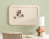 Rectangular beige fabric pinboard with rounded light wooden frame hanging on pale green wall with white diamond pattern, displaying three photos; below is a white desk with a gold lamp, pink flowers, and an open book.